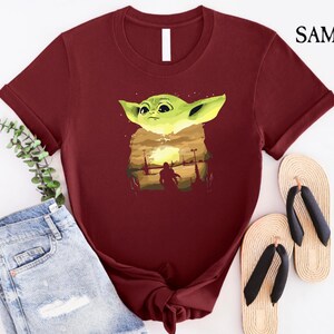 May include: A maroon t-shirt with a graphic of a green alien character and a sunset scene. The shirt is styled with blue denim shorts and beige flip-flops. The text "SAMART" is visible in the upper right corner.