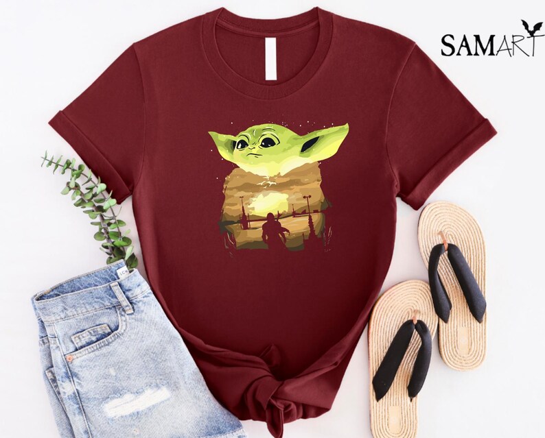 May include: A maroon t-shirt with a graphic of a green alien character and a sunset scene. The shirt is styled with blue denim shorts and beige flip-flops. The text "SAMART" is visible in the upper right corner.