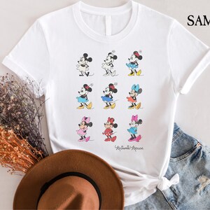 May include: White t-shirt with a graphic of nine different Minnie Mouse characters in various poses. The text "Minnie Mouse" is printed at the bottom of the shirt.
