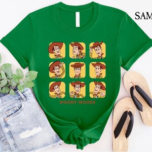 以下が含まれることがあります： トイストーリーのウッディの漫画の画像がプリントされた緑色のTシャツ。画像は、ウッディがさまざまな表情をグリッドパターンで示しています。画像の下には「ウッディムード」というテキストが印刷されています。
