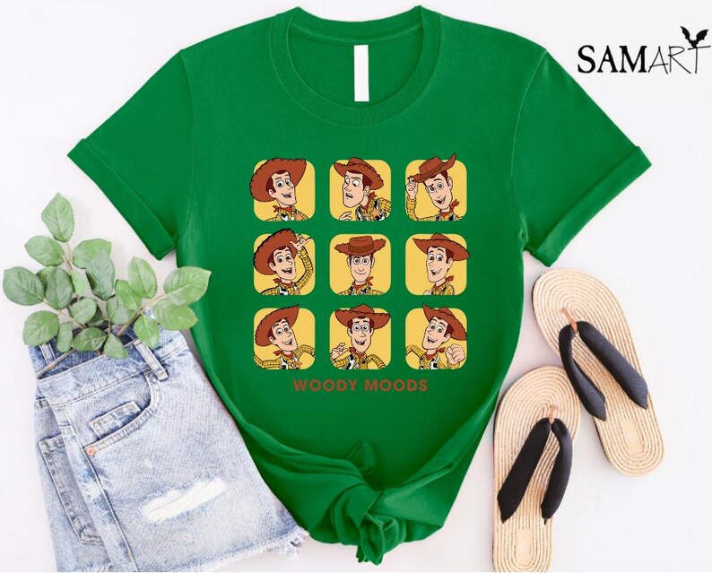 以下が含まれることがあります： トイストーリーのウッディの漫画の画像がプリントされた緑色のTシャツ。画像は、ウッディがさまざまな表情をグリッドパターンで示しています。画像の下には「ウッディムード」というテキストが印刷されています。