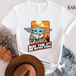 May include: White t-shirt with a cartoon image of a green alien character from Star Wars. The text "Star Wars Day May the 4th Be With You" is printed on the shirt.