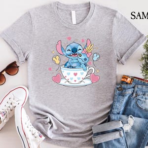 May include: A light gray t-shirt featuring a cartoon illustration of Stitch and Angel in a teacup, surrounded by hearts and balloons. The shirt is paired with blue jeans, white sneakers, and sunglasses.