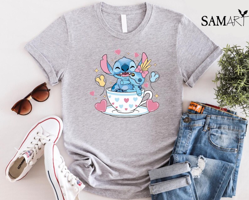 May include: A light gray t-shirt featuring a cartoon illustration of Stitch and Angel in a teacup, surrounded by hearts and balloons. The shirt is paired with blue jeans, white sneakers, and sunglasses.