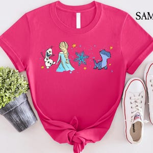 May include: A hot pink t-shirt featuring a cartoon design of characters from the movie Frozen, including Elsa, Olaf, and Bruni. The shirt is paired with white sneakers. The brand name "SAMART" is printed in the upper right corner.