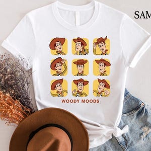 以下が含まれることがあります： トイストーリーのウッディの漫画の画像がプリントされた白いTシャツ。画像は、ウッディがさまざまな表情をグリッドパターンで示しています。画像の下には「ウッディムード」というテキストが印刷されています。