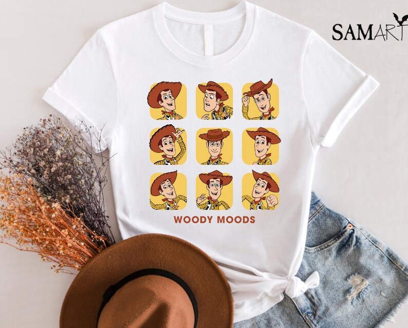 以下が含まれることがあります： トイストーリーのウッディの漫画の画像がプリントされた白いTシャツ。画像は、ウッディがさまざまな表情をグリッドパターンで示しています。画像の下には「ウッディムード」というテキストが印刷されています。