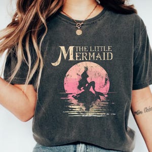 May include: A black t-shirt with a vintage-style graphic of Ariel, the Little Mermaid, sitting on a rock in the ocean. The text "The Little Mermaid" is printed above the graphic.
