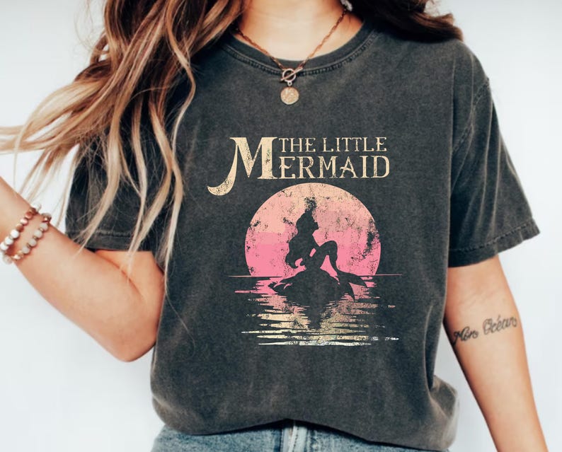 May include: A black t-shirt with a vintage-style graphic of Ariel, the Little Mermaid, sitting on a rock in the ocean. The text "The Little Mermaid" is printed above the graphic.