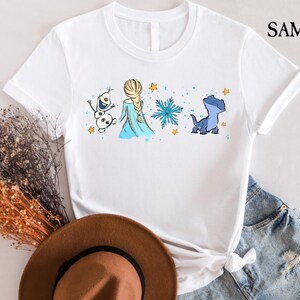 May include: White t-shirt featuring a cartoon illustration of Elsa, Olaf, and a blue dragon-like creature from the movie Frozen. The design includes a snowflake and stars. The shirt has the text "SAMART" on the upper right.