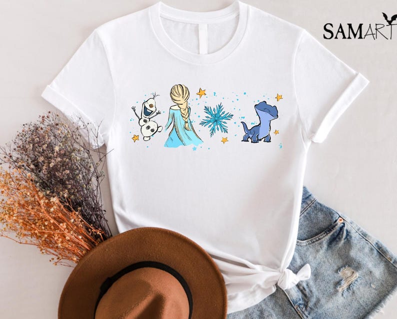 May include: White t-shirt featuring a cartoon illustration of Elsa, Olaf, and a blue dragon-like creature from the movie Frozen. The design includes a snowflake and stars. The shirt has the text "SAMART" on the upper right.