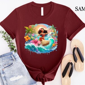 Pode incluir: Camiseta bord&ocirc; com uma ilustra&ccedil;&atilde;o vibrante de uma mulher dan&ccedil;ando em uma onda, vestindo uma roupa vermelha e branca. O design inclui flores tropicais, palmeiras e um p&ocirc;r do sol. A camiseta tem gola redonda e mangas curtas.