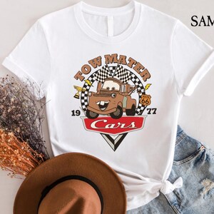 May include: White t-shirt with a vintage-style graphic of Tow Mater from the Disney Pixar movie Cars. The graphic features Tow Mater with the text "Tow Mater" above him and "Cars" below him. The graphic also includes a checkered flag and Route 66 sign.