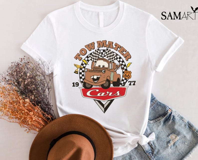 May include: White t-shirt with a vintage-style graphic of Tow Mater from the Disney Pixar movie Cars. The graphic features Tow Mater with the text "Tow Mater" above him and "Cars" below him. The graphic also includes a checkered flag and Route 66 sign.