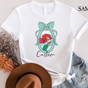 May include: White t-shirt featuring a cartoon illustration of a mermaid with red hair and a green tail, framed by a teal bow and oval border. The word "Custom" is written below the image.