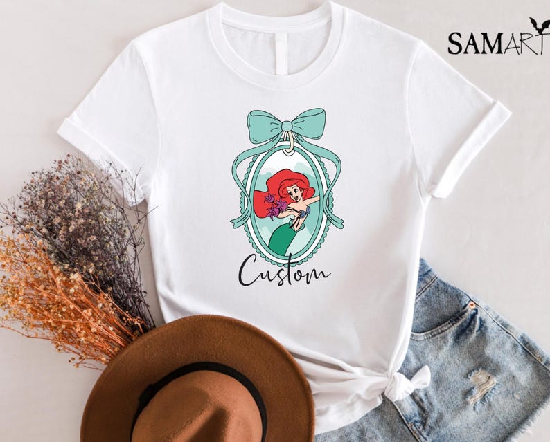 May include: White t-shirt featuring a cartoon illustration of a mermaid with red hair and a green tail, framed by a teal bow and oval border. The word "Custom" is written below the image.