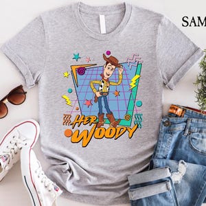 以下が含まれることがあります： トイストーリーのウッディのレトロなグラフィックがプリントされたグレーのTシャツ。グラフィックは、カラフルな幾何学模様と「Her Woody」の文字が書かれた青と緑のグリッドの中にウッディが描かれています。
