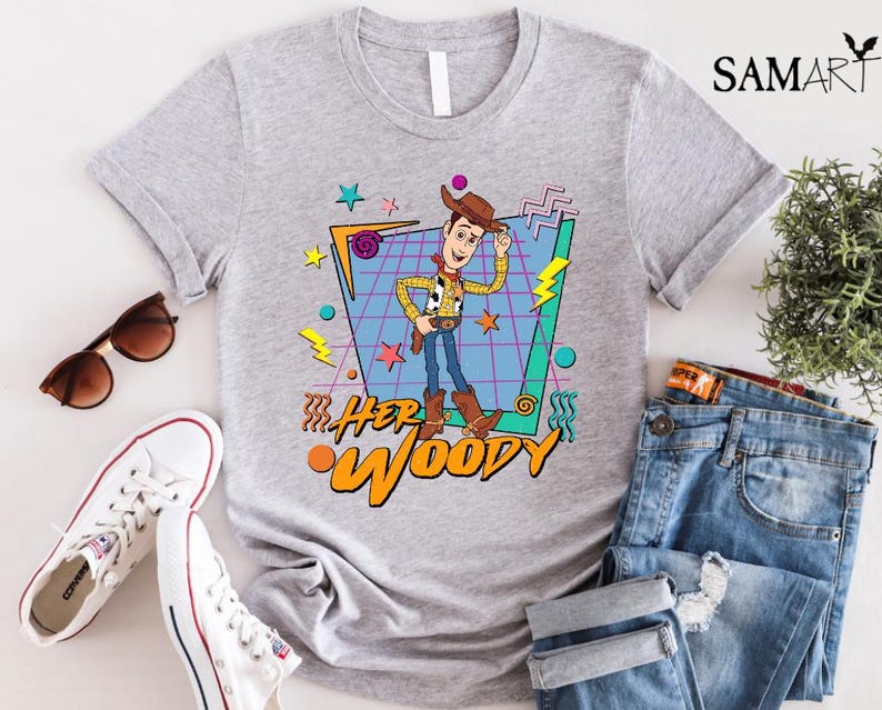 以下が含まれることがあります： トイストーリーのウッディのレトロなグラフィックがプリントされたグレーのTシャツ。グラフィックは、カラフルな幾何学模様と「Her Woody」の文字が書かれた青と緑のグリッドの中にウッディが描かれています。