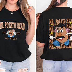 Könnte beinhalten: Schwarzes T-Shirt mit einem braunen und weißen Aufdruck von Mr. Potato Head aus Toy Story. Der Text "MR. POTATO HEAD" ist in braunen Buchstaben über dem Aufdruck gedruckt. Der Text "EST. 1995" ist in braunen Buchstaben unter dem Aufdruck gedruckt. Der Text "TOY STORY" ist in braunen Buchstaben unter dem Text "EST. 1995" gedruckt.