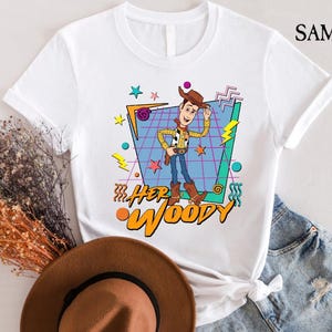 以下が含まれることがあります： トイストーリーのウッディのレトロなグラフィックがプリントされた白いTシャツ。グラフィックは、青と紫のグリッドにカラフルな幾何学模様と「Her Woody」の文字が描かれています。