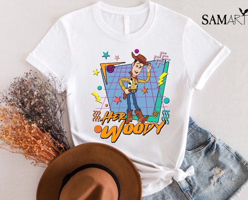 以下が含まれることがあります： トイストーリーのウッディのレトロなグラフィックがプリントされた白いTシャツ。グラフィックは、青と紫のグリッドにカラフルな幾何学模様と「Her Woody」の文字が描かれています。