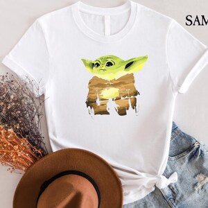 May include: White t-shirt with a cartoon character in green and yellow, set against a brown and yellow landscape. The shirt has short sleeves and the text "SAMART" is printed on the upper right side.