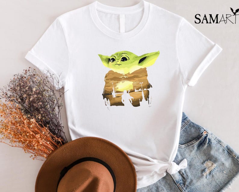 May include: White t-shirt with a cartoon character in green and yellow, set against a brown and yellow landscape. The shirt has short sleeves and the text "SAMART" is printed on the upper right side.
