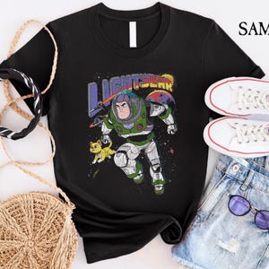 May include: Black t-shirt featuring a graphic of Buzz Lightyear from the Toy Story franchise. The graphic depicts Buzz in his space ranger suit with a yellow cat in the background. The text "LIGHTYEAR" is printed above Buzz.