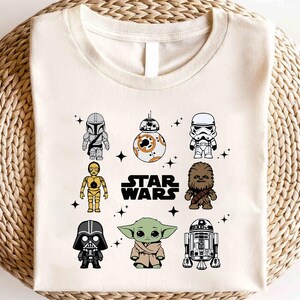 K&ouml;nnte beinhalten: Cremefarbenes T-Shirt mit Cartoon-Illustrationen von Star Wars-Charakteren, darunter der Mandalorianer, BB-8, ein Sturmtruppler, C-3PO, Chewbacca, Darth Vader, Grogu und R2-D2. Die Worte "STAR WARS" sind in Schwarz gedruckt.