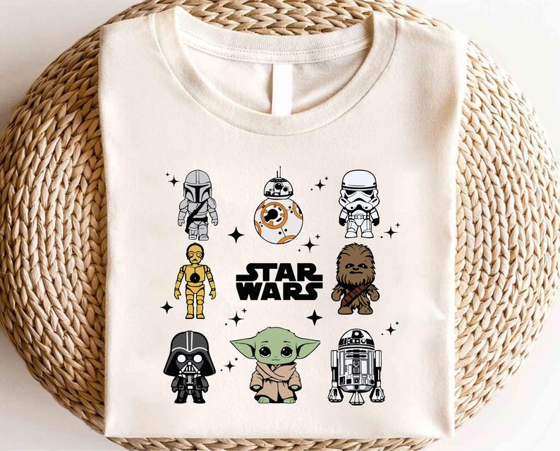 K&ouml;nnte beinhalten: Cremefarbenes T-Shirt mit Cartoon-Illustrationen von Star Wars-Charakteren, darunter der Mandalorianer, BB-8, ein Sturmtruppler, C-3PO, Chewbacca, Darth Vader, Grogu und R2-D2. Die Worte "STAR WARS" sind in Schwarz gedruckt.