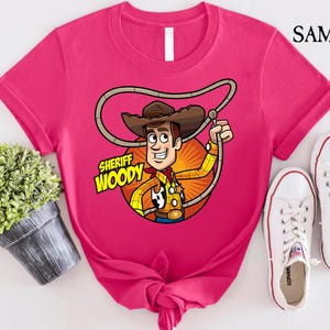 Pode incluir: Camiseta rosa com uma imagem de desenho animado do Woody do Toy Story. A imagem mostra o Woody usando seu distintivo de xerife e seu chap&eacute;u. O texto "Sheriff Woody" est&aacute; impresso na camiseta.