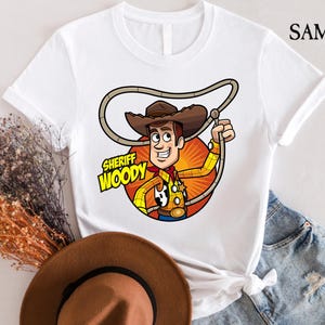 Pode incluir: Camiseta branca com uma imagem de desenho animado do Woody do Toy Story. Woody est&aacute; usando seu chap&eacute;u de cowboy e colete caracter&iacute;sticos. O texto "Sheriff Woody" est&aacute; impresso na camiseta.