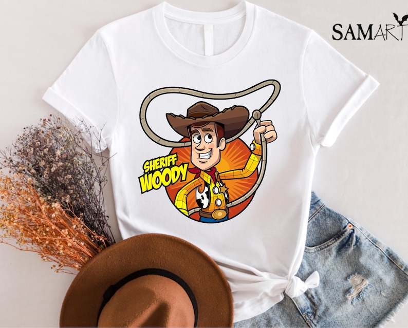 Pode incluir: Camiseta branca com uma imagem de desenho animado do Woody do Toy Story. Woody est&aacute; usando seu chap&eacute;u de cowboy e colete caracter&iacute;sticos. O texto "Sheriff Woody" est&aacute; impresso na camiseta.