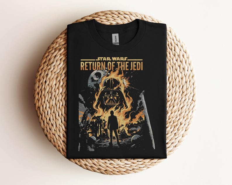 Pode incluir: T-shirt preta com um design de Star Wars. O gr&aacute;fico inclui o capacete de Darth Vader em chamas, o texto "STAR WARS RETURN OF THE JEDI" e uma silhueta. A t-shirt &eacute; apresentada numa superf&iacute;cie tecida.