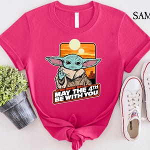 May include: Pink t-shirt with a cartoon image of a green alien character with big ears and a brown robe. The text "Star Wars Day May the 4th Be With You" is printed on the shirt.
