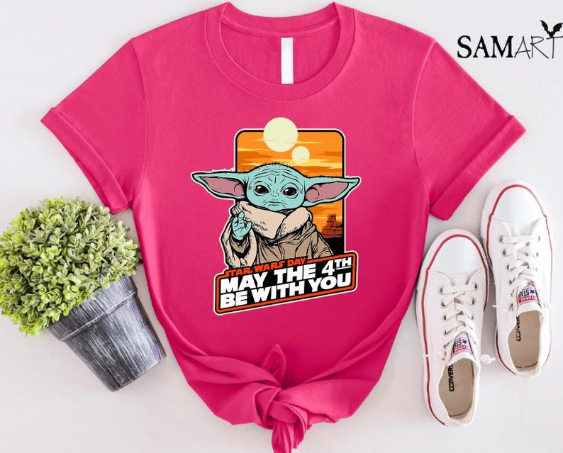 May include: Pink t-shirt with a cartoon image of a green alien character with big ears and a brown robe. The text "Star Wars Day May the 4th Be With You" is printed on the shirt.