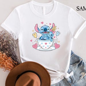 May include: White t-shirt featuring a cartoon illustration of Stitch and Angel in a teacup, surrounded by hearts and stars. The design includes the text "SAMART" in the upper right corner. The shirt is paired with denim shorts.