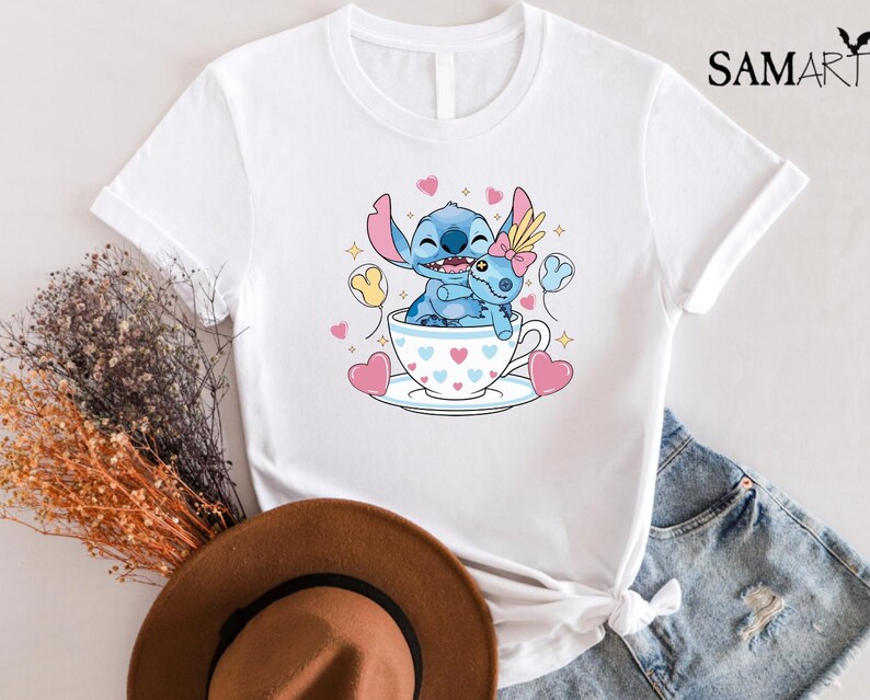 May include: White t-shirt featuring a cartoon illustration of Stitch and Angel in a teacup, surrounded by hearts and stars. The design includes the text "SAMART" in the upper right corner. The shirt is paired with denim shorts.