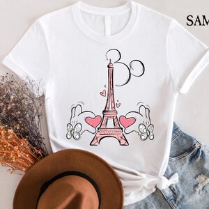 Puede incluir: Camiseta blanca con una ilustraci&oacute;n rosa y negra de la Torre Eiffel con manos de Mickey Mouse sosteniendo corazones a cada lado. La ilustraci&oacute;n es de estilo de dibujos animados.