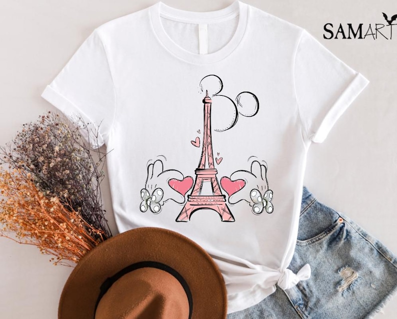 Puede incluir: Camiseta blanca con una ilustraci&oacute;n rosa y negra de la Torre Eiffel con manos de Mickey Mouse sosteniendo corazones a cada lado. La ilustraci&oacute;n es de estilo de dibujos animados.