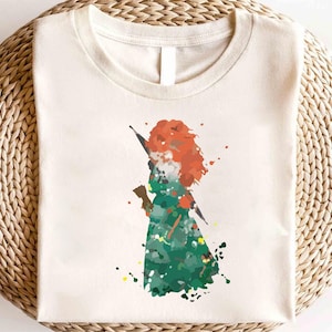 Puede incluir: Una camiseta blanca con una ilustración al estilo acuarela de una mujer pelirroja sosteniendo una espada. La mujer lleva un vestido verde y está rodeada de salpicaduras de pintura.