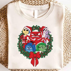 May include: A white t-shirt with a Christmas wreath design featuring the characters from the movie Inside Out. The characters are Joy, Sadness, Anger, Fear, and Disgust. The wreath is decorated with red and green holly.