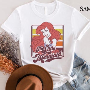 May include: White t-shirt with a retro-style graphic of Ariel, the Little Mermaid, with the text "The Little Mermaid" in a colorful, vintage font.