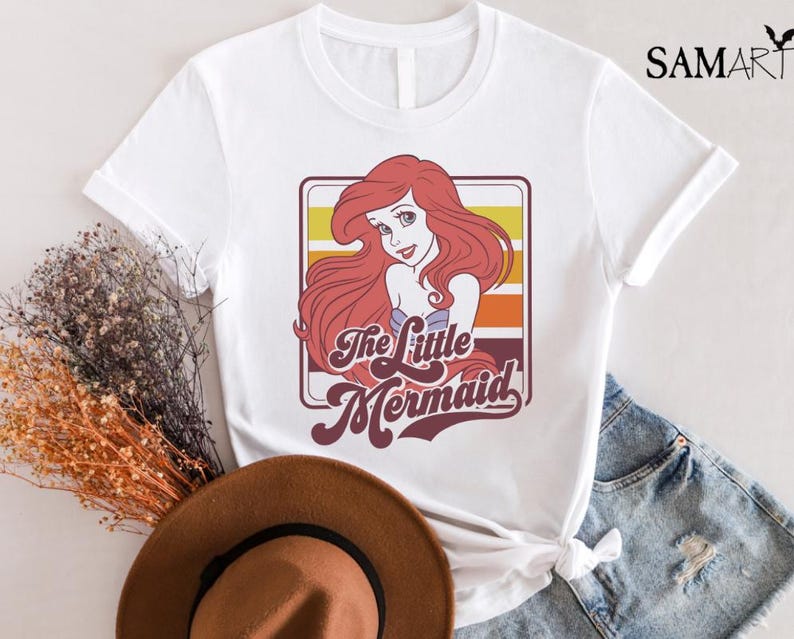 May include: White t-shirt with a retro-style graphic of Ariel, the Little Mermaid, with the text "The Little Mermaid" in a colorful, vintage font.