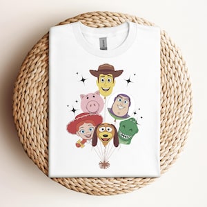 Op de afbeelding: Wit T-shirt met een kleurrijke afbeelding van Toy Story-personages als ballonnen. Het ontwerp bevat Woody, Hamm, Jessie, Buzz Lightyear, Rex en Slinky Dog. Het shirt wordt gepresenteerd op een geweven, cirkelvormig oppervlak.