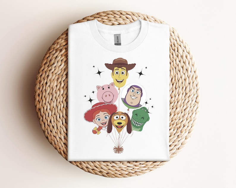 Op de afbeelding: Wit T-shirt met een kleurrijke afbeelding van Toy Story-personages als ballonnen. Het ontwerp bevat Woody, Hamm, Jessie, Buzz Lightyear, Rex en Slinky Dog. Het shirt wordt gepresenteerd op een geweven, cirkelvormig oppervlak.