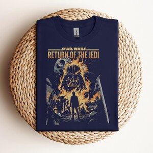 Pode incluir: Camiseta azul marinho com o t&iacute;tulo do filme "Star Wars: O Retorno de Jedi". O design mostra o capacete de Darth Vader em chamas, uma silhueta e a Estrela da Morte. A camiseta est&aacute; dobrada sobre uma superf&iacute;cie tecida.