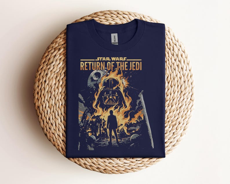Pode incluir: Camiseta azul marinho com o t&iacute;tulo do filme "Star Wars: O Retorno de Jedi". O design mostra o capacete de Darth Vader em chamas, uma silhueta e a Estrela da Morte. A camiseta est&aacute; dobrada sobre uma superf&iacute;cie tecida.