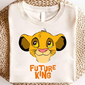May include: A white t-shirt with a cartoon image of Simba from The Lion King. The image is in yellow, orange, and black. The text "FUTURE KING" is printed in orange below the image.
