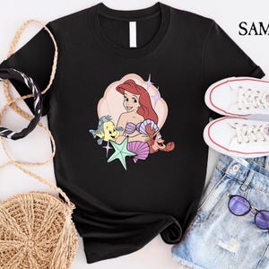 May include: Black t-shirt featuring a colorful graphic of Ariel from The Little Mermaid, with Flounder, Sebastian, and a starfish. The design is set against a light pink and cream background. The shirt is styled with white sneakers and denim shorts.